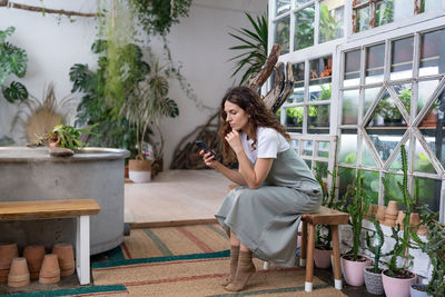 Serious young gardener woman owner holding smartphone reading media news, sitting in home garden