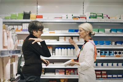 Side view of senior female customer explaining to owner standing by medicine rack at pharmacy store