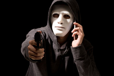 Criminal in mask using mobile phone while aiming with handgun against black background