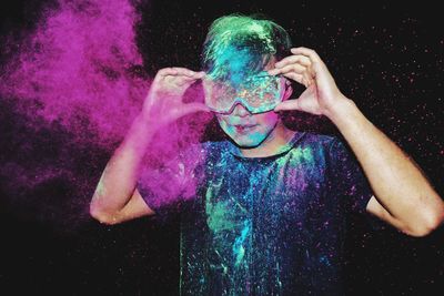 Young man with powder paint adjusting sunglasses against black background