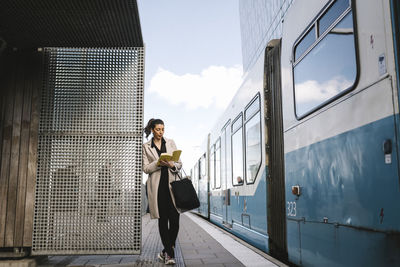 Businesswoman reading book while leaning at metal wall by train at railroad station