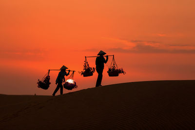 Silhouette men carrying potted plants in baskets on bamboo pole on landscape against orange sky