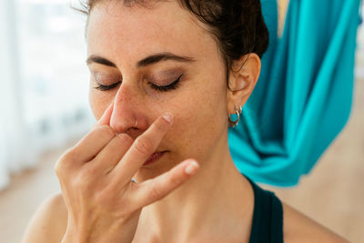 Calm female with closed eyes doing breathing exercise while sitting in light studio with colorful hanging swings for aerial yoga