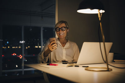 Smiling businesswoman using smart phone while sitting with laptop at illuminated desk in coworking space