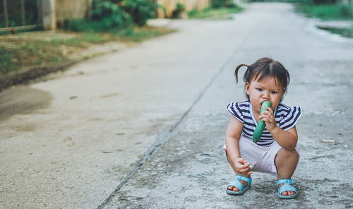 Portrait of baby girl eating cucumber while crouching on footpath