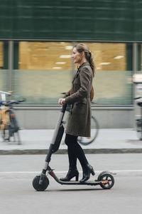 Full length side view of businesswoman riding electric push scooter on street in city