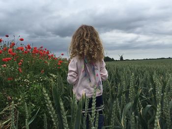 Rear view of girl standing amidst plants on field against cloudy sky