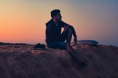 Man sitting on rock against sky during sunset