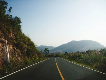 Empty road along trees and mountains against clear sky