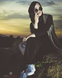 Young woman sitting on sunglasses against sky during sunset