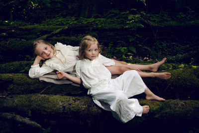 Two girls sisters in white dresses lie on logs in the moss in the forest