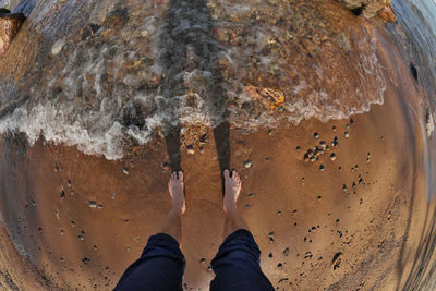 First person fisheye perspective of man's feet in shallow water with waves rolling into sandy beach