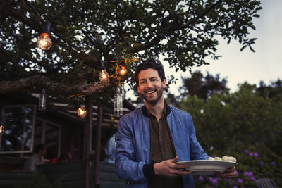 Portrait of happy man holding plate while standing at yard during dinner party