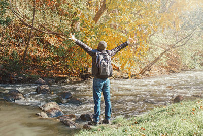 Full length of man with arms raised standing by stream at forest