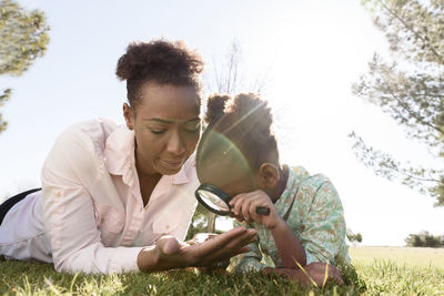 Daughter looking at mother's hand with magnifying glass while lying on grassy field in park during sunny day