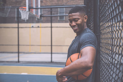 Smiling young man holding basketball by chainlink fence