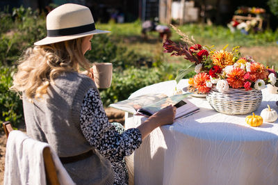 A woman in a hat with a brim flips through a book and drinks a hot drink at a table 