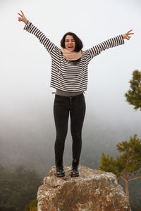 Full length of carefree young woman with arms raised standing on rock during foggy weather