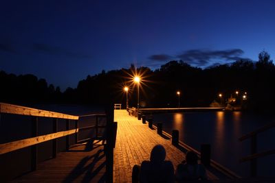 Rear view of people on illuminated pier against sky at dusk