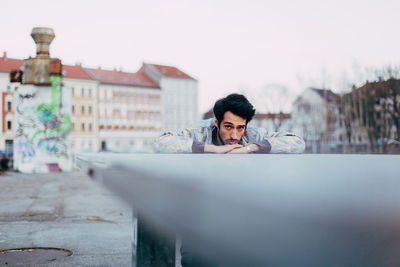 Portrait of young man leaning on railing against sky in city