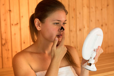 Beautiful young woman applying facial mask on face while sitting in sauna