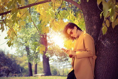 Side view of mid adult woman using while standing by tree trunk