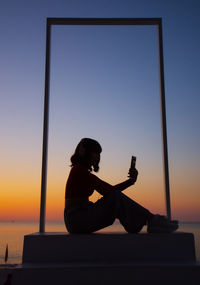 Silhouette woman sitting on sea against sky during sunset