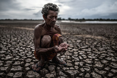 Shirtless man holding falling water while crouching on cracked land against sky