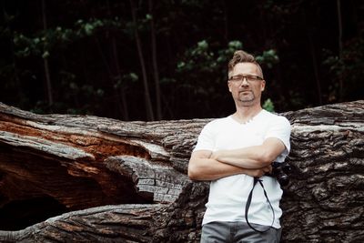 Portrait of mature man holding camera while standing by log in forest