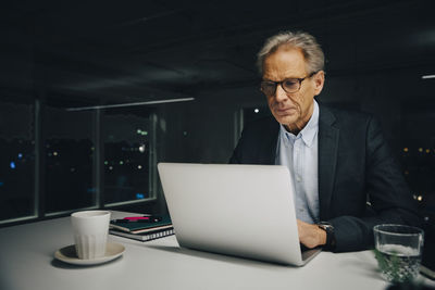 Confident senior businessman using laptop while sitting at illuminated desk in creative office