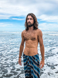 Shirtless man looking away while standing at beach against sky