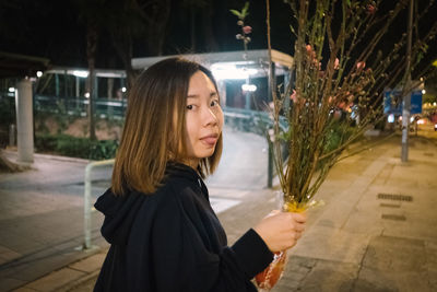 Portrait of woman holding bouquet while standing on footpath at night