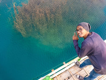 High angle portrait of young man gesturing while standing on pier over sea