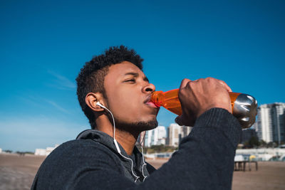 Young man drinking water from bottle while listening music against sky