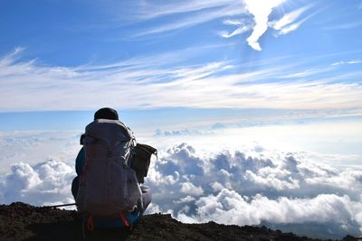 Rear view of man looking at clouds while sitting on mountain against sky