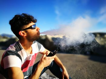 Young man smoking electronic cigarette while sitting on field against sky