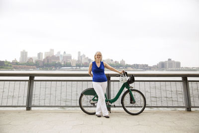 Portrait of happy mature woman standing with bicycle on promenade in city against clear sky