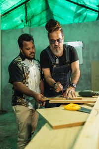 Serious multiracial male colleagues with ruler measuring wooden plank while standing at table with saw in professional joinery with special equipment