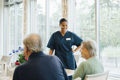 Smiling young female nurse looking at senior woman sitting by man at dining table in elderly nursing home