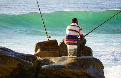 Rear view of man fishing on rock by sea