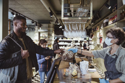 Smiling male customer collecting order from female store owner from deli store during covid-19