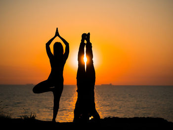 Silhouette couple exercising at beach against sky during sunset