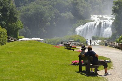 Couple sitting on bench at park against waterfall