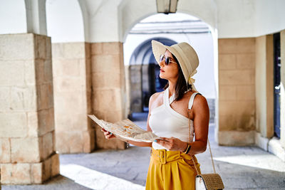 Thoughtful female tourist in hat and sunglasses reading paper map while standing on street during summer vacation and orientating in city