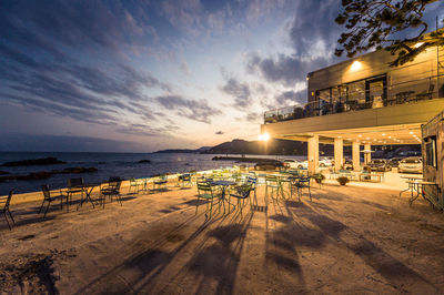 Chairs and tables at illuminated restaurant by sea against cloudy sky at dusk