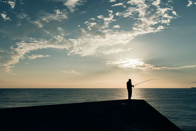 Silhouette man fishing in sea while standing on pier against sky during sunset