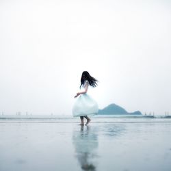 Full length of woman wearing white dress while spinning at beach against sky