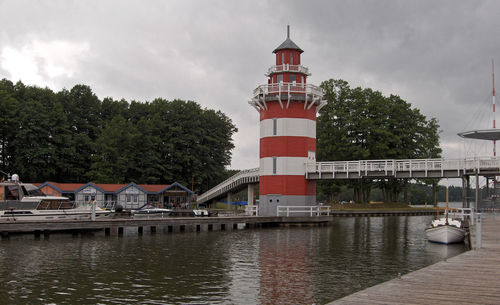 Lighthouse by river and buildings against sky