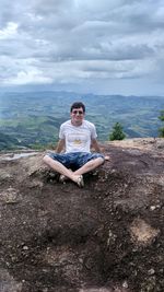 Smiling young man sitting on cliff against cloudy sky