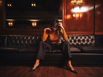Fashionable pensive woman in stylish clothes sitting wide spread apart legs on black leather couch with head down in cabinet